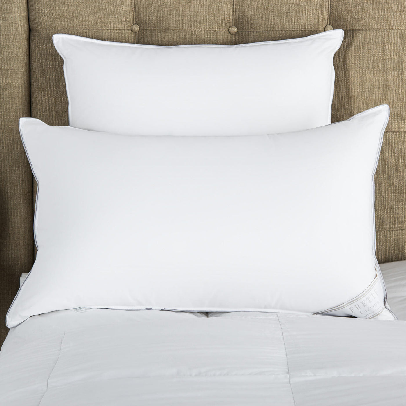 Two Frette Cortina Down Pillows are stacked against a beige upholstered headboard on a neatly made bed with a white comforter.