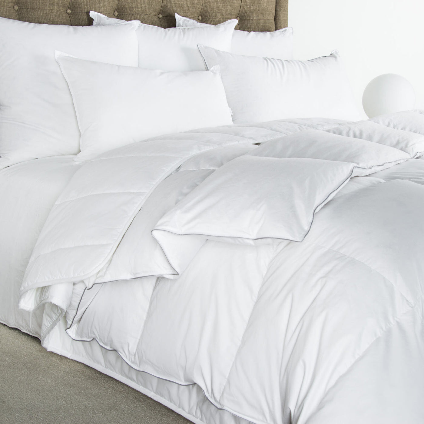 A neatly made bed with the Frette Cortina Down Duvet Insert in a cotton sateen finish, multiple white pillows, and a beige upholstered headboard.
