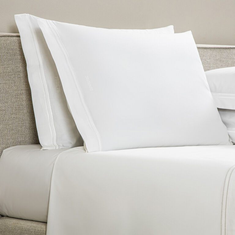 Two Frette Classic pillowcases by Frette in white rest against a beige upholstered headboard atop a neatly made bed with cotton percale sheets, adding a touch of luxury bedding.