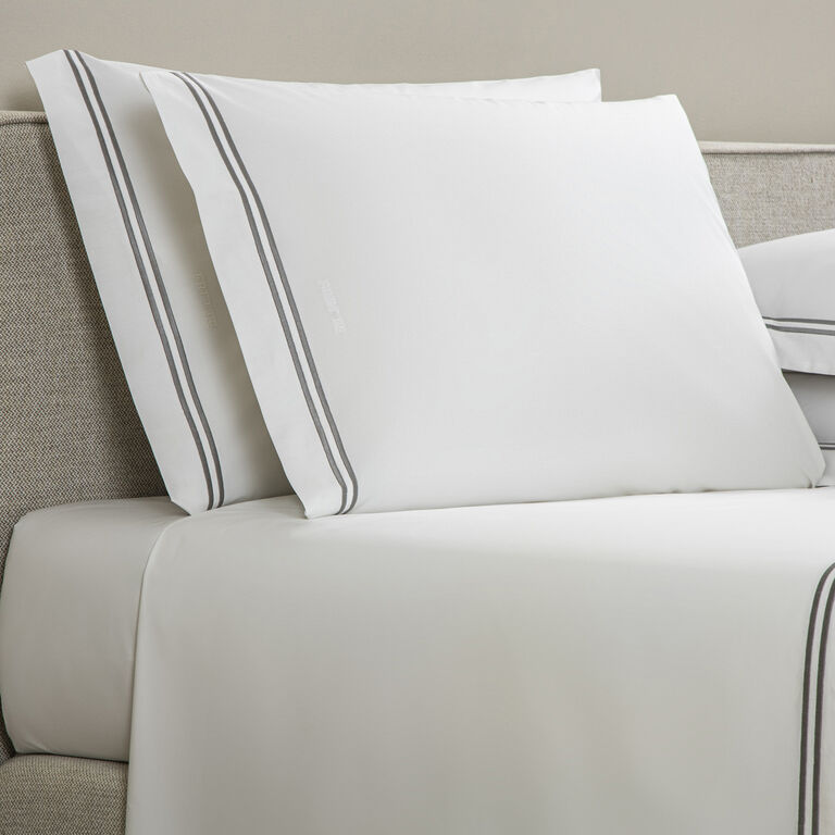 Two white Frette Classic | Pillowcase Set pillowcases with grey double-line borders rest against a beige headboard on a neatly made bed, offering a touch of luxury bedding from Frette.