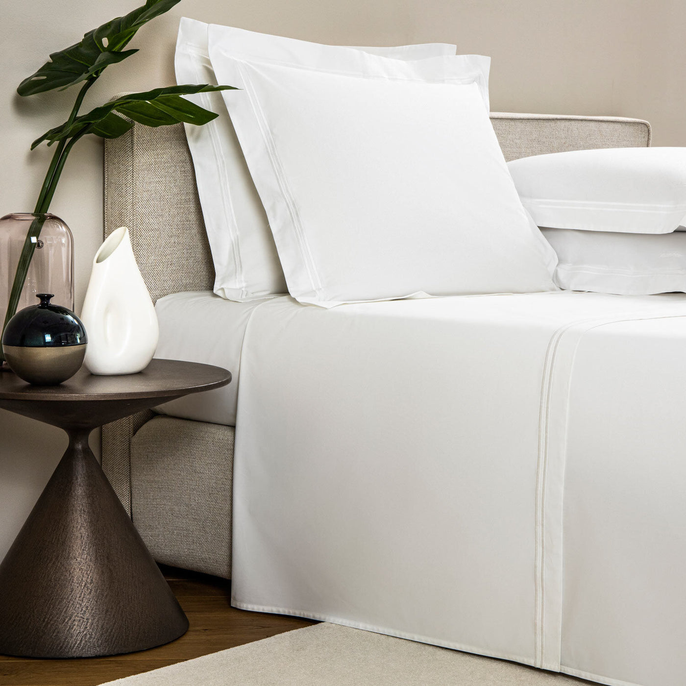 A neatly made bed with white sheets and pillows from the Frette Classic | Sheet Set by Frette sits beside a modern round side table displaying decorative vases and a green leafy plant.
