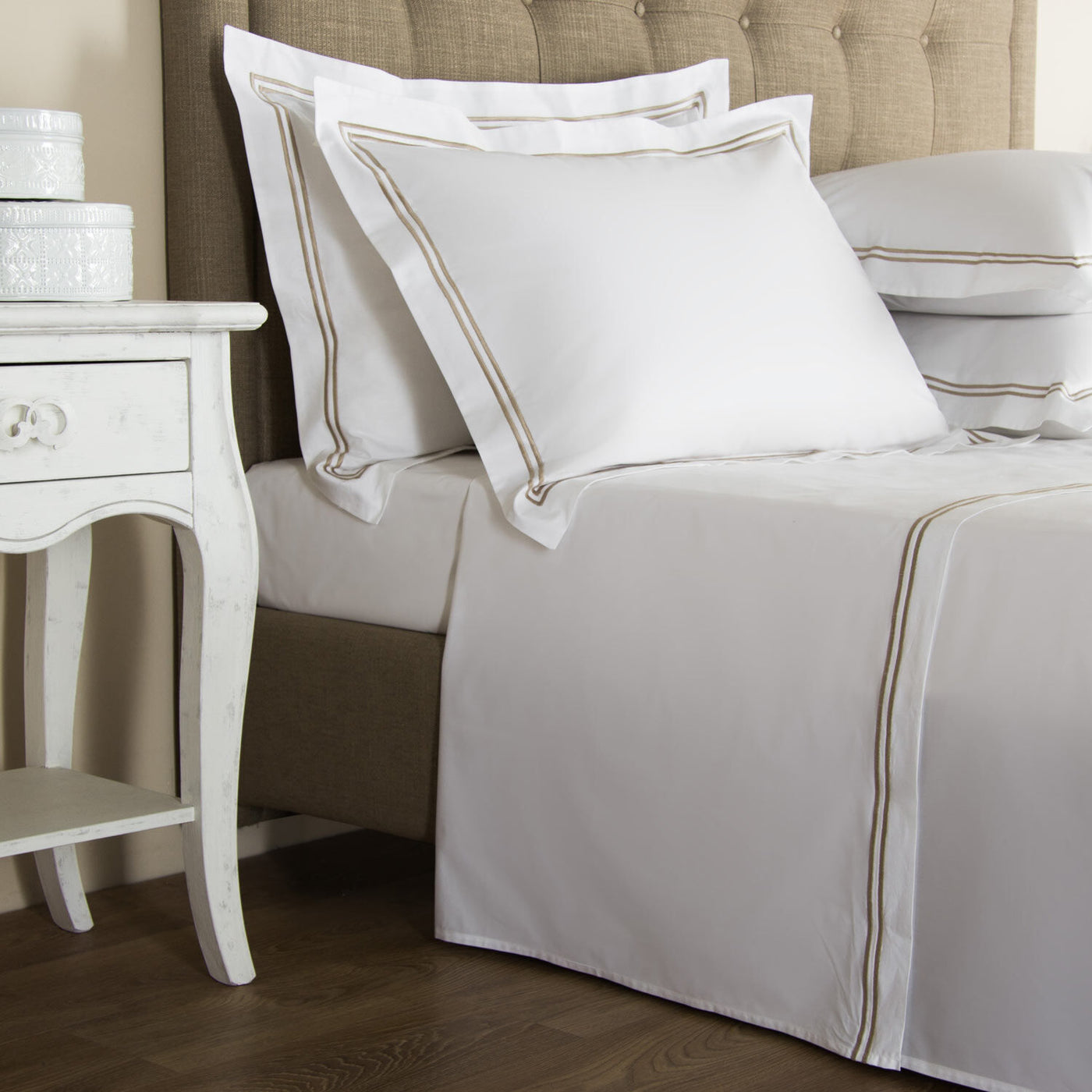 White bed with tan trim and luxury bedding, Frette Classic pillow shams by Frette, matching pillowcases, a tufted beige headboard, and a white nightstand with stacked boxes, on a wooden floor.