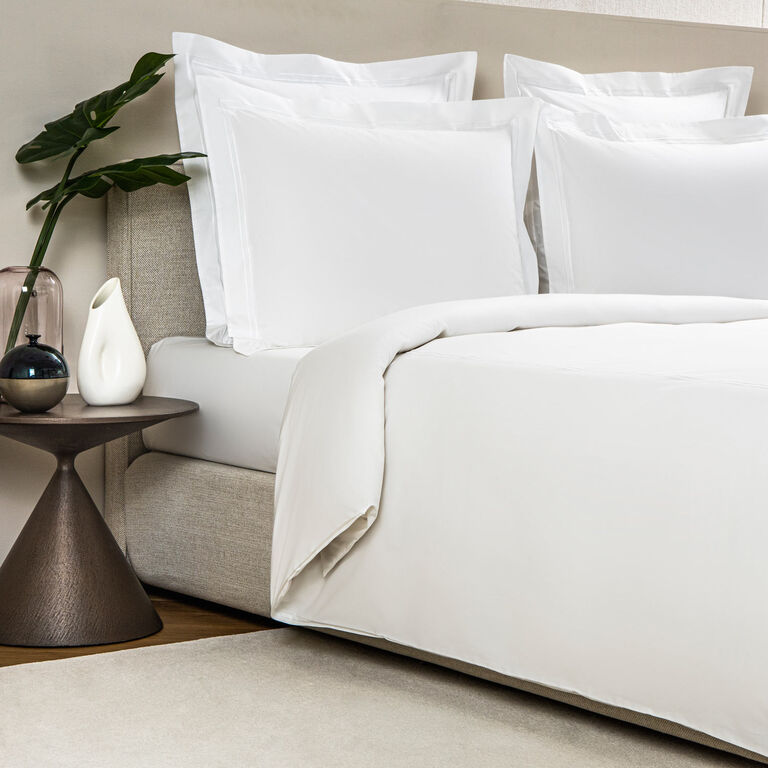 A neatly made bed features the Frette Classic | Duvet Cover by Frette in white, paired with pillows. Beside it is a round side table topped with a glass vase holding a green leaf and a white decorative object.