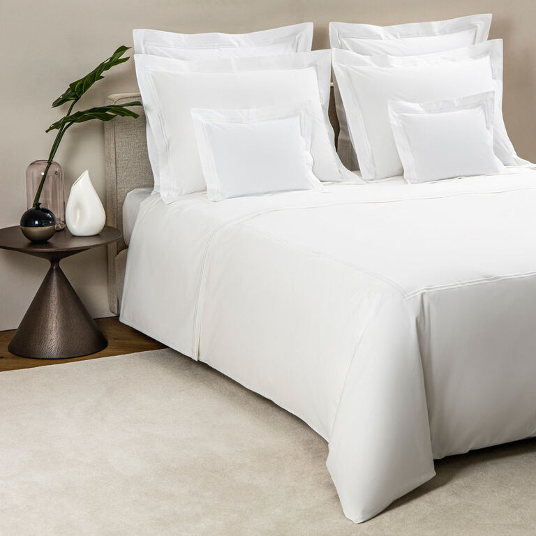 A neatly made bed features the Frette Classic | Duvet Cover by Frette and multiple pillows, placed beside a round side table holding a vase, a green leafy plant, and a white decorative object.
