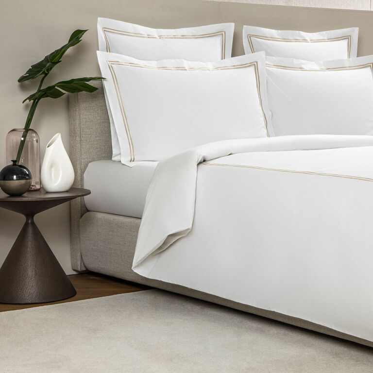 A neatly made bed features the Frette Classic Duvet Cover—white cotton percale with gold trim—beside a modern nightstand topped with two vases and a green leafy plant.