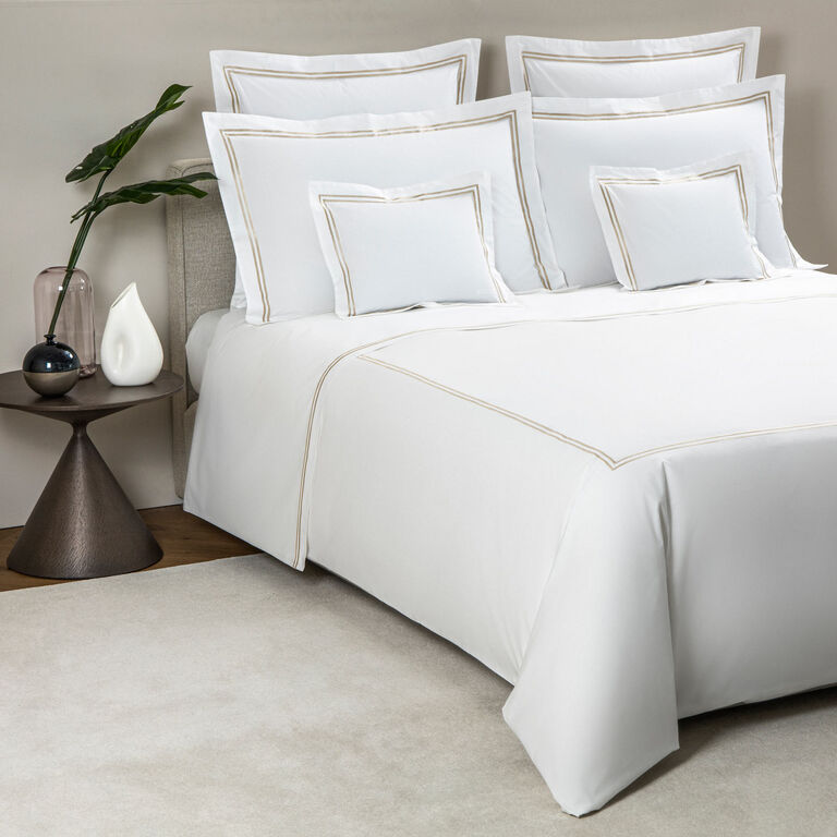 A neatly made bed features the Frette Classic | Duvet Cover by Frette in white with gold trim, complemented by matching pillows. A round side table with a plant and decor sits beside the luxury bedding set.