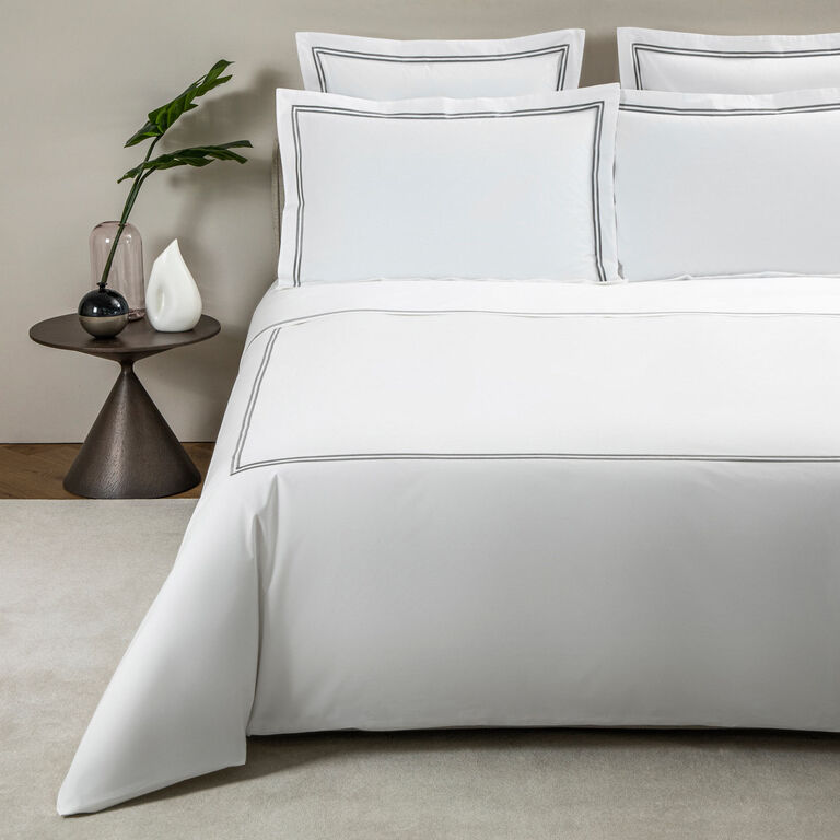 A neatly made bed with the Frette Classic | Duvet Cover, white cotton percale with a thin grey border by Frette, beside a round table holding a vase, decor accents, and green foliage.