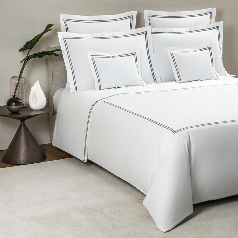 A neatly made bed with Frette’s Classic Duvet Cover—white cotton percale with grey trim—paired with five pillows, a bedside table with a vase, decorative accents, and a light-colored rug.