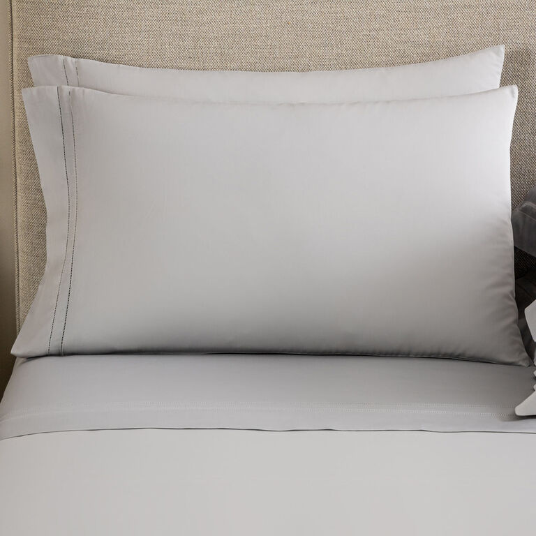 Two light gray Frette Doppio Ajour pillowcases are stacked against a beige fabric headboard, paired with matching light gray bedsheets.