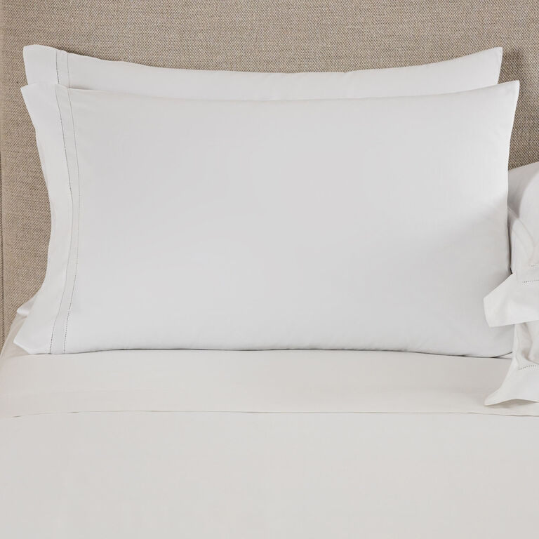 A neatly made bed features two stacked white pillows in Frette Doppio Ajour | Pillowcase Set, paired with crisp cotton sateen shams, against a beige upholstered headboard.