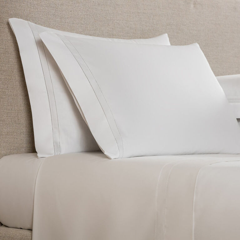 Two white Frette Doppio Ajour pillowcases are stacked against a beige upholstered headboard on a neatly made bed with matching white sheets.
