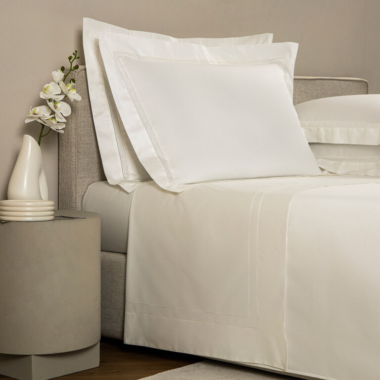 A neatly made bed with luxury bedding features the Frette Doppio Ajour | Sheet Set in cotton sateen, next to a round nightstand holding a vase of white flowers and stacked coasters.