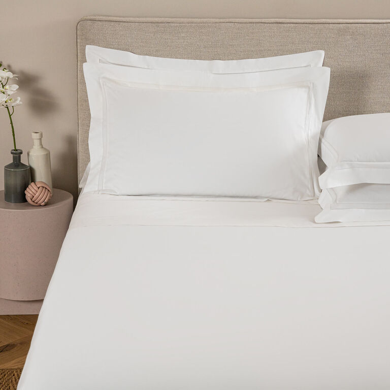 A neatly made bed with white sheets and Frette Doppio Ajour Pillow Shams sits beside a round bedside table adorned with decorative vases and a small white orchid.