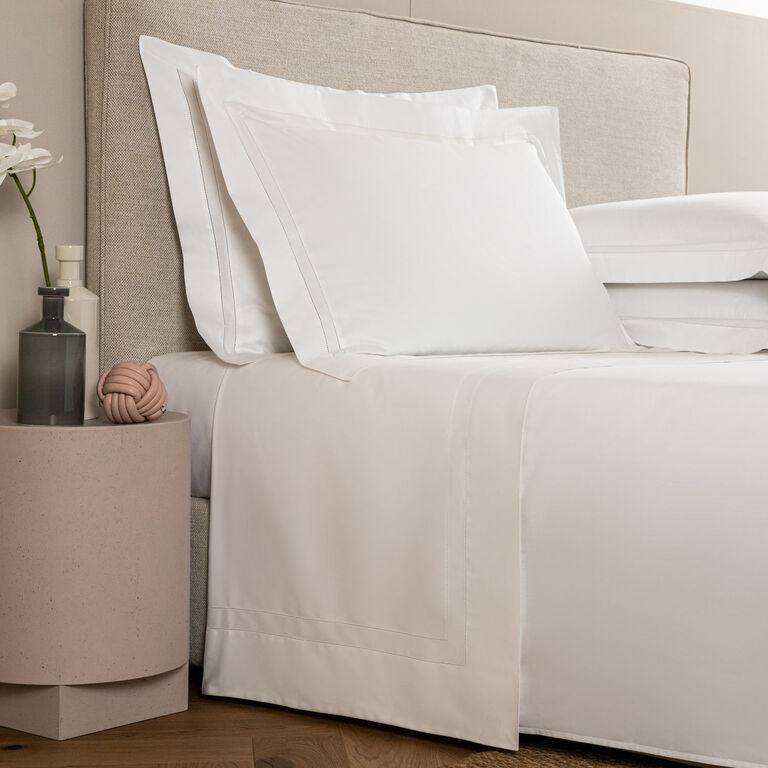 A neatly made bed with white Frette Doppio Ajour | Sheet Set linens and pillows sits beside a round pink nightstand topped with a vase, decorative object, and flowers—a scene of modern luxury.