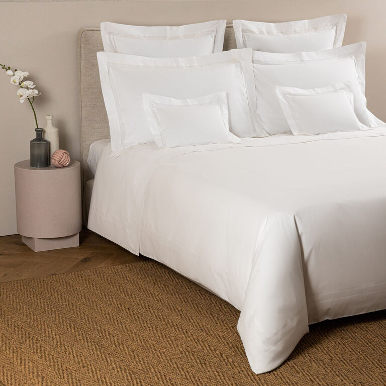 A neatly made bed with white linens and pillows features the Frette Doppio Ajour Duvet Cover in smooth cotton sateen, beside a round nightstand with a vase of white flowers and decor, atop a woven rug.