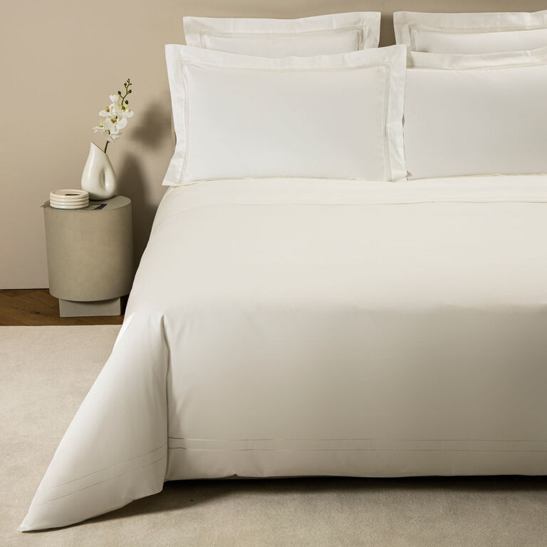 A neatly made bed with white cotton sateen bedding and pillows features the Frette Doppio Ajour Duvet Cover with double open hemstitch, beside a small round table holding a white vase of flowers and stacked dishes.