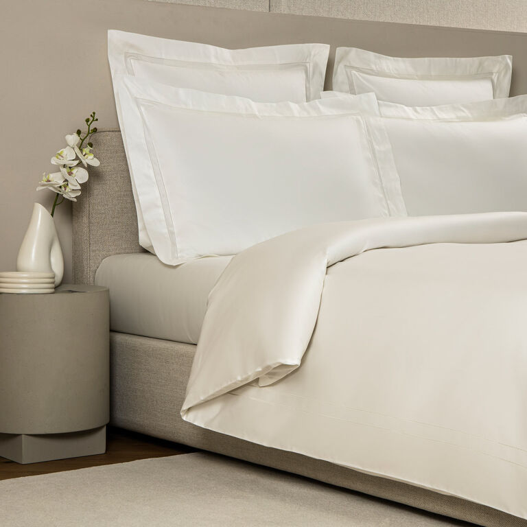 A neatly made bed features the Frette Doppio Ajour Duvet Cover and pillowcases, beside a gray nightstand with a white vase holding an orchid.