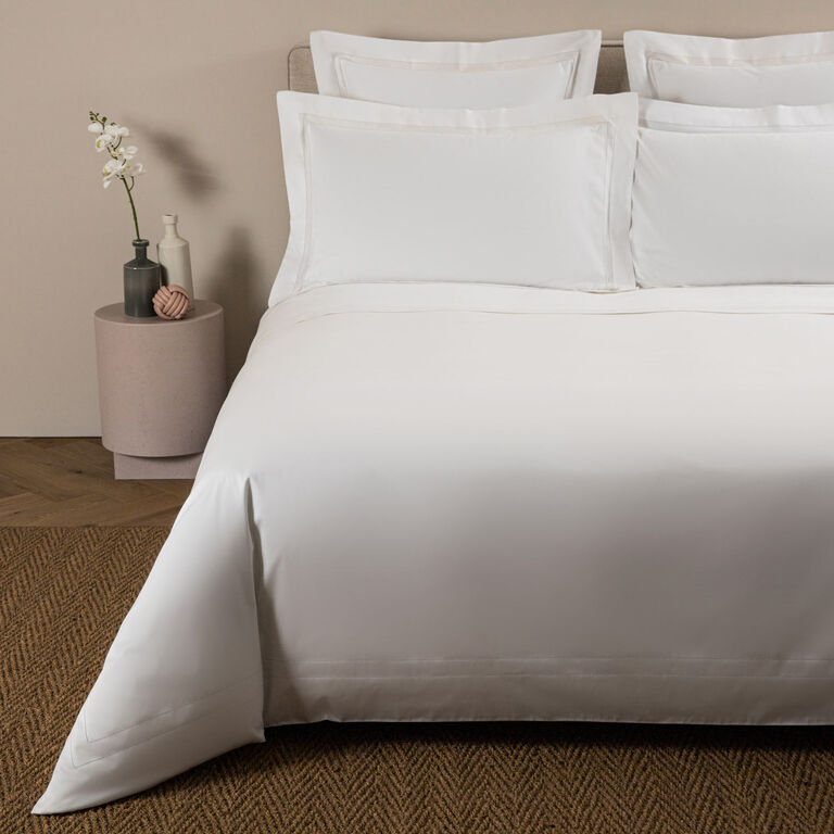 A bed made with white cotton sateen bedding and pillows is topped with the Frette Doppio Ajour Duvet Cover, featuring a double open hemstitch, and sits next to a round table with a small plant on a woven rug.