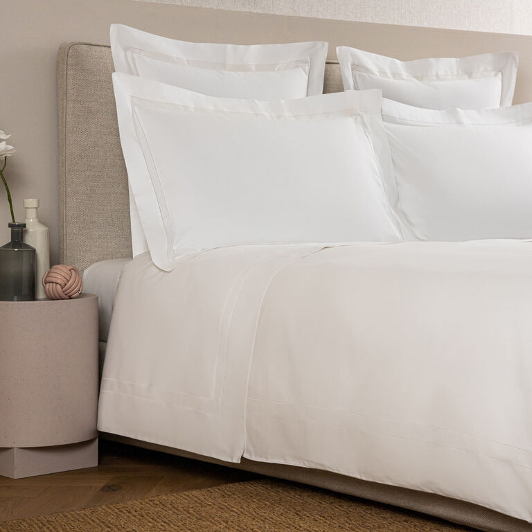 A neatly made bed features white pillows and a Frette Doppio Ajour Duvet Cover in soft cotton sateen, beside a round table with a vase holding a white flower, decorative objects, and a dark bottle.