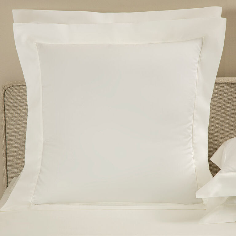 The Grace | Pillow Sham by Frette, crafted from long-staple cotton sateen, features elegant shadow stitching and rests against a beige upholstered headboard, adding refined detail and luxury to the bed.
