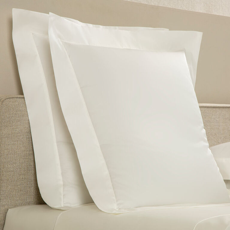 Two large pillows with Frette’s Grace Pillow Shams in smooth long-staple cotton sateen are propped against a beige upholstered headboard on a neatly made bed.