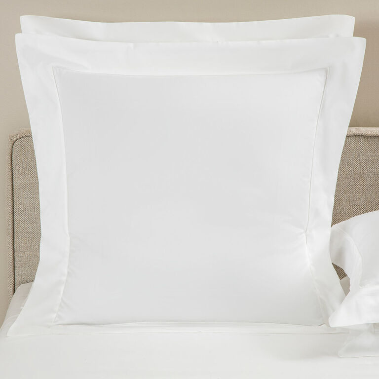 A Frette Grace Pillow Sham in white, featuring elegant shadow stitching and crafted from long-staple cotton sateen, is propped against a beige headboard on a bed with crisp white sheets.