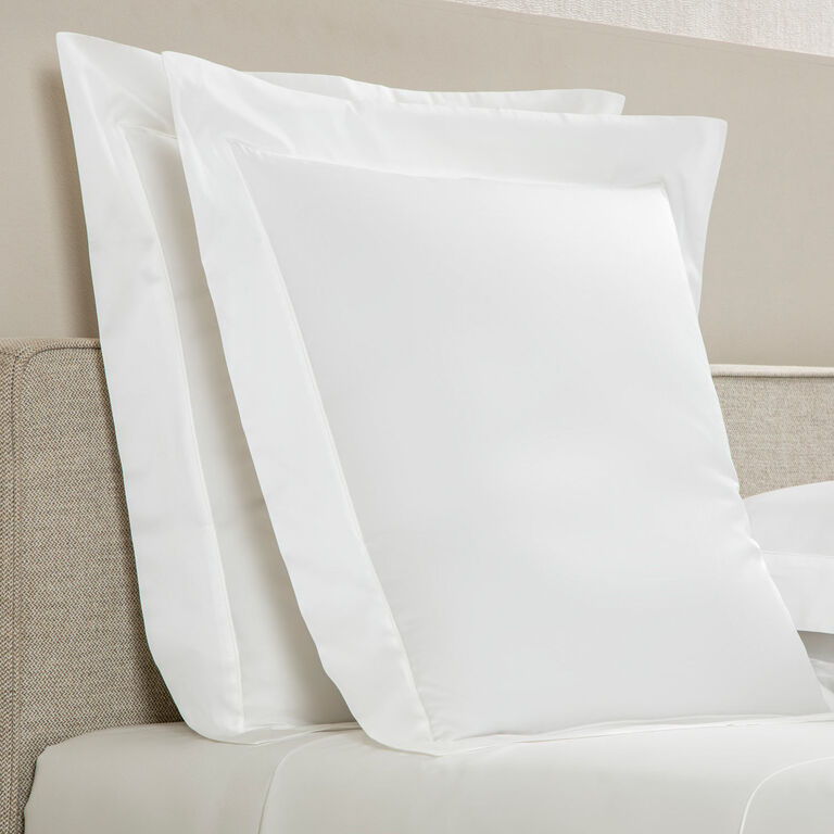 Two large white pillows with Frette’s Grace Pillow Shams are propped against a beige fabric headboard on a neatly made bed.