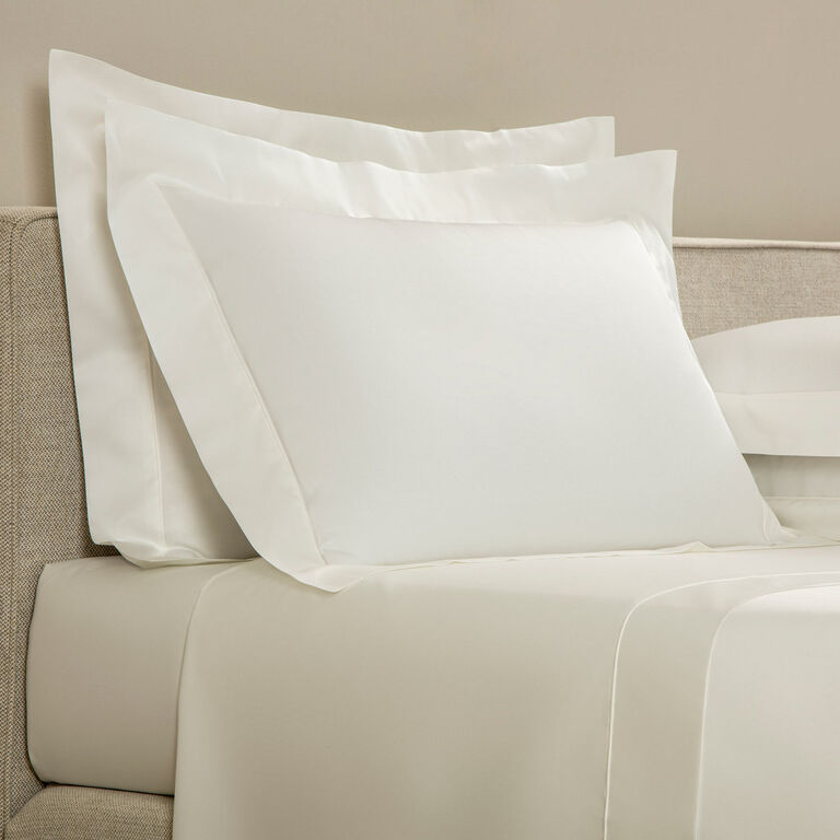 A neatly made bed features Frette's Grace Pillow Shams of white long-staple cotton sateen with an elegant shadow stitch, resting against a beige upholstered headboard.