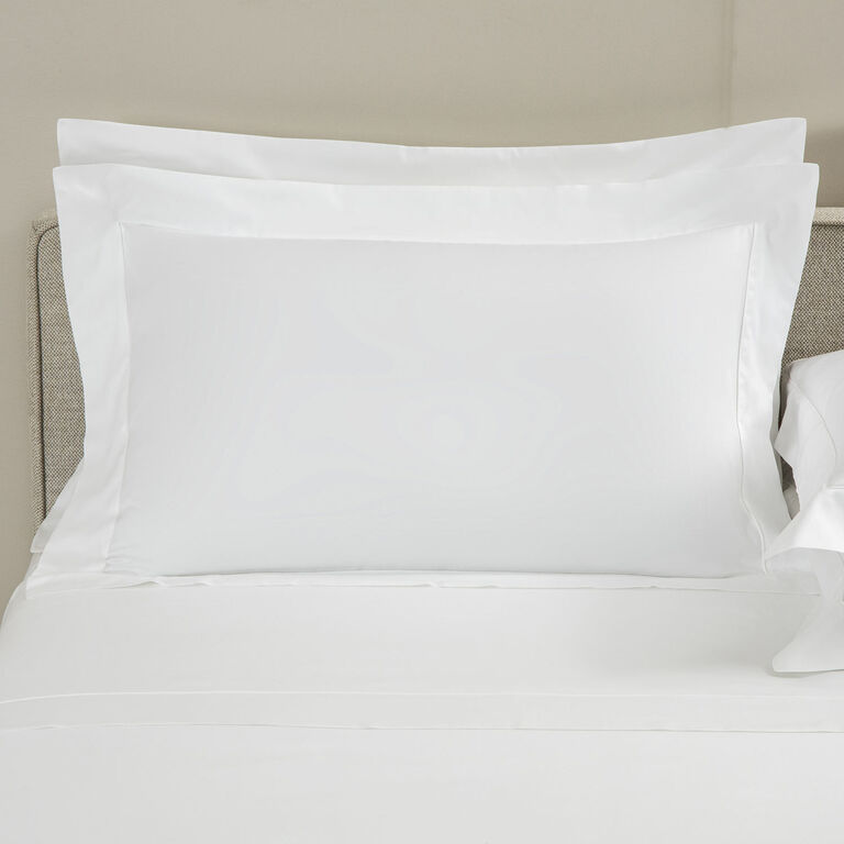 The Grace | Pillow Sham by Frette, with an elegant shadow stitch and wide flange, is displayed neatly on a bed against a light-colored headboard and wall.