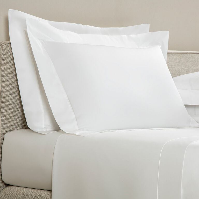 Two white Frette Grace pillow shams are arranged on a beige upholstered bed, complemented by matching white long-staple cotton sateen sheets and bedding.