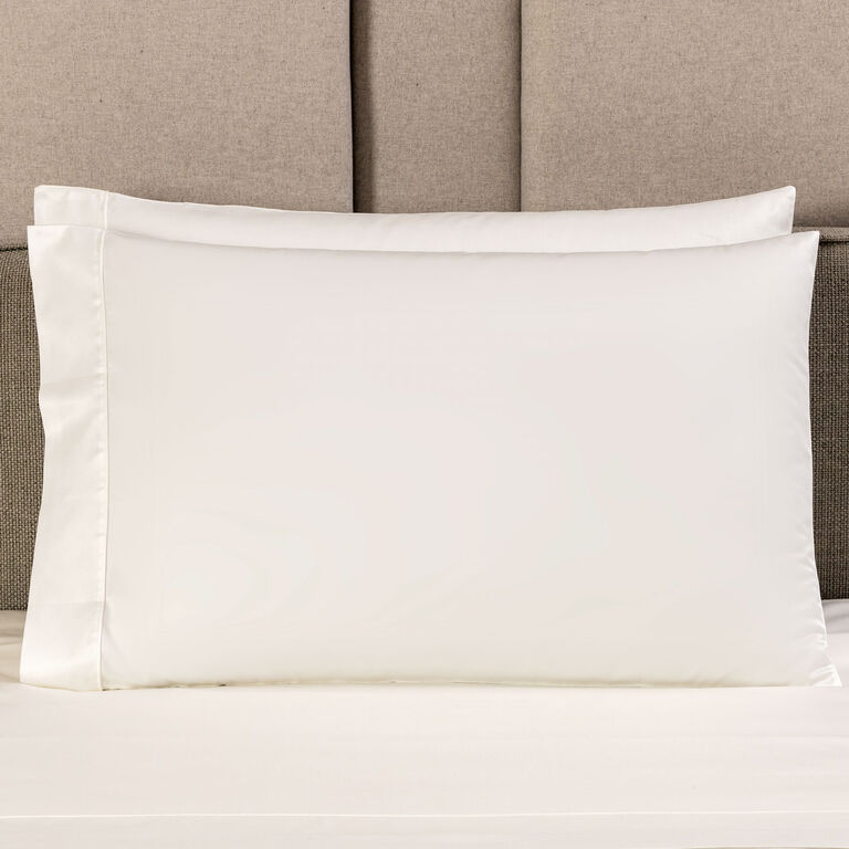 A white pillow with a smooth Grace | Pillowcase Set by Frette rests on a bed, with a beige upholstered headboard in the background.