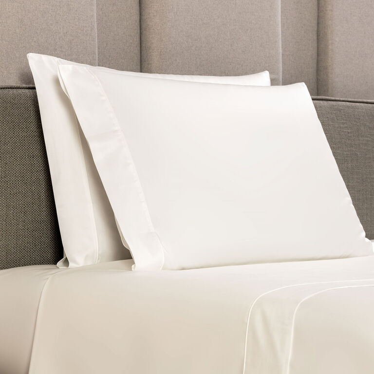 Two white pillows with Frette's Grace pillowcase set are arranged on a neatly made bed with a light gray upholstered headboard.