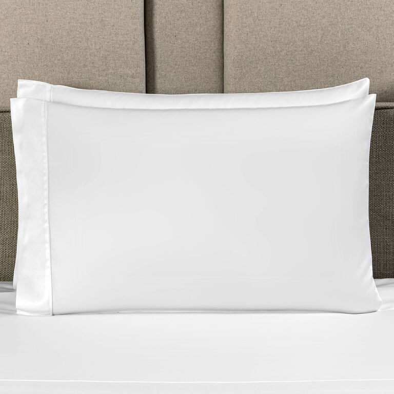 A white pillow in the Grace | Pillowcase Set by Frette rests against a beige upholstered headboard on a neatly made bed.