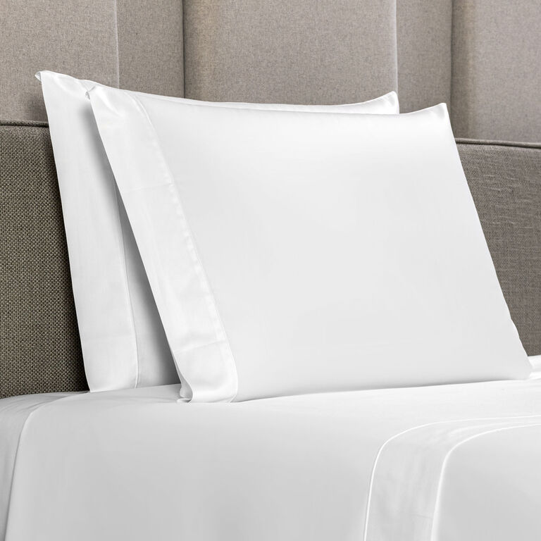 Two Grace | Pillowcase Set by Frette, crafted from long-staple cotton sateen, rest on pillows atop crisp white sheets with a padded, light gray headboard in the background.