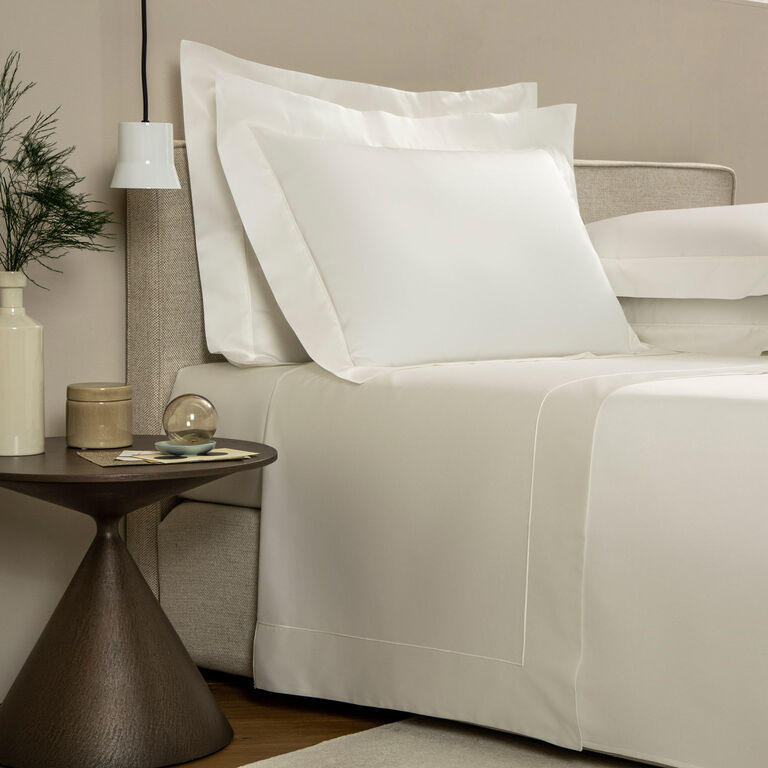A neatly made bed features the Grace Sheet Set by Frette in white cotton sateen, paired with a small round side table holding a glass sphere, a vase with greenery, and a hanging white pendant light.