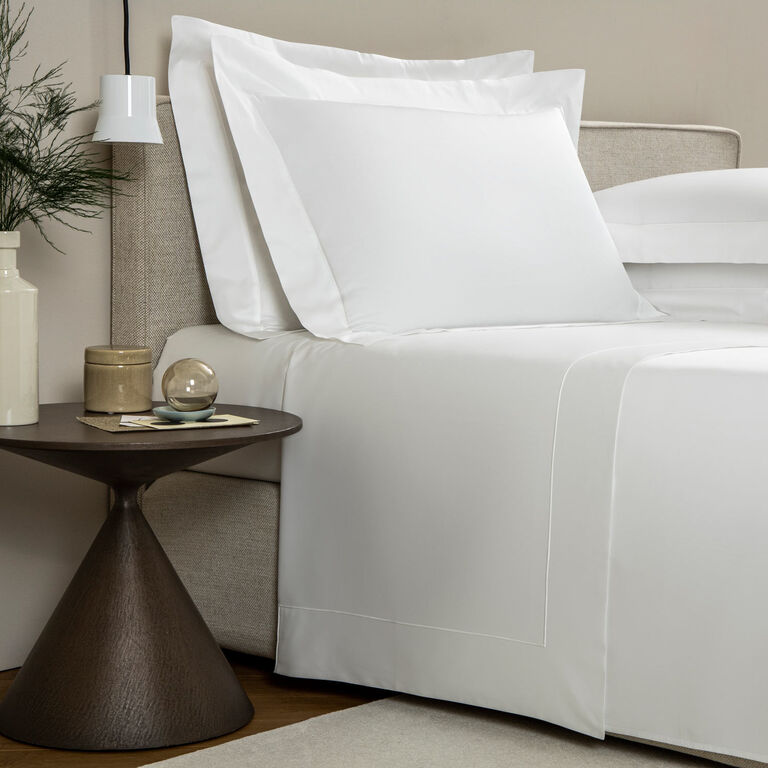A neatly made bed with Frette’s Grace | Sheet Set in crisp white long-staple cotton sits beside a modern round side table with decor in a simple, contemporary bedroom.