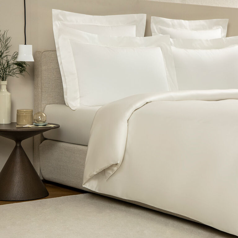 A neatly made bed with the Frette Grace duvet cover in white cotton sateen and pillows stands beside a small round side table holding a lamp, books, and decorative items.