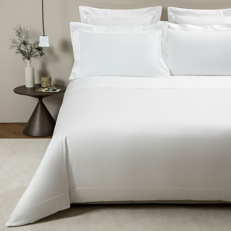 A neatly made bed features the Grace | Duvet Cover by Frette in soft white cotton sateen, accented with pillows, beside a small round table holding a vase with greenery, a candle, and small items.