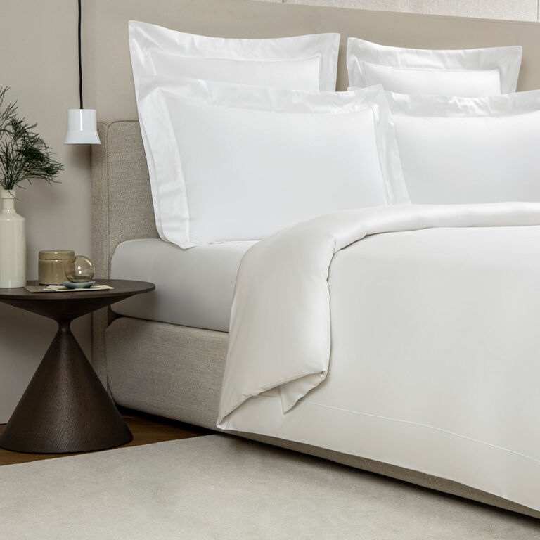 A neatly made bed features the Grace Duvet Cover by Frette in elegant white cotton, accompanied by a small round side table topped with a vase, plant, and lamp.