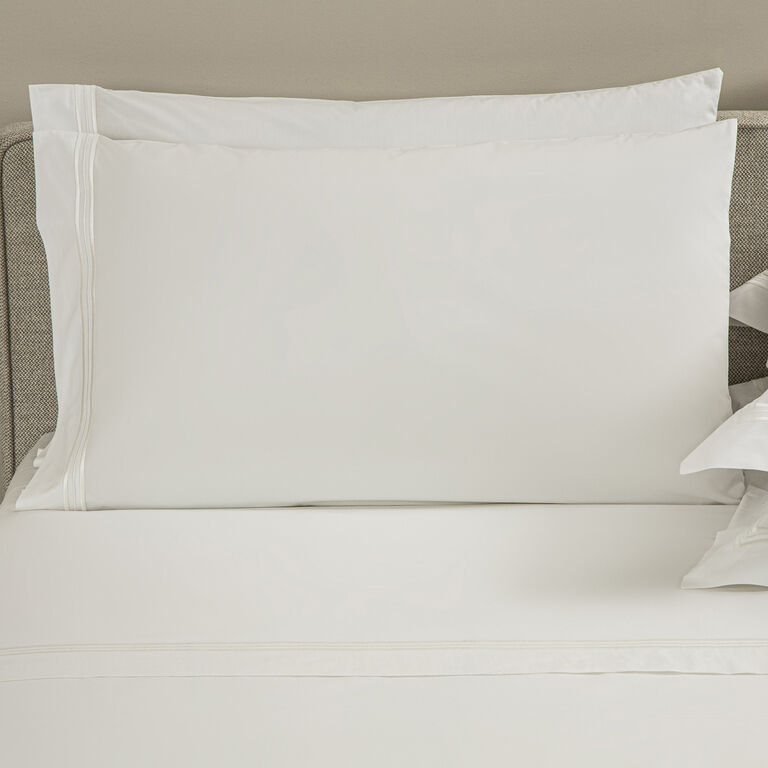 A neatly made bed features Frette’s Triplo Bourdon Pillowcase Set in white cotton poplin, with a large and a smaller pillow against a beige headboard, both accented by subtle three-line embroidery.