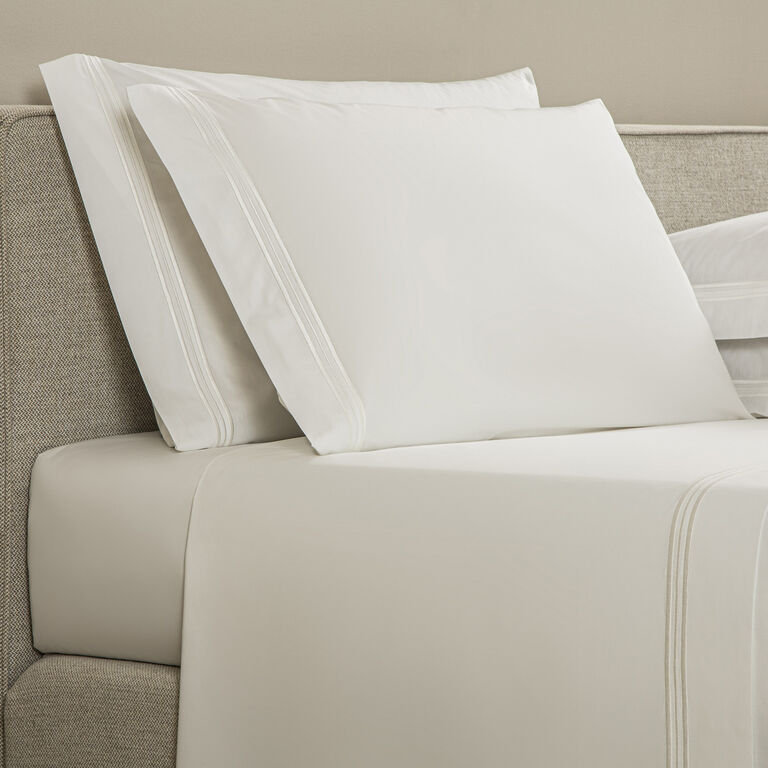 A neatly made bed with Frette’s Triplo Bourdon Pillowcase Set in white cotton poplin, featuring three-line embroidery, complements a beige upholstered headboard.