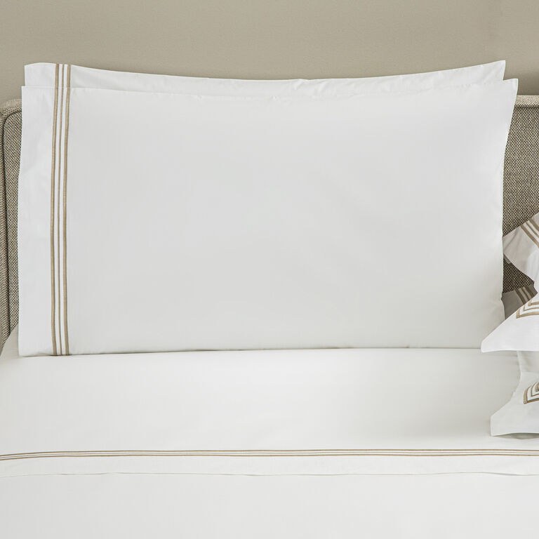 The Triplo Bourdon Pillowcase Set by Frette features white pillowcases with beige and brown striped borders and elegant three-line embroidery, displayed on a matching bedspread against a light beige headboard.