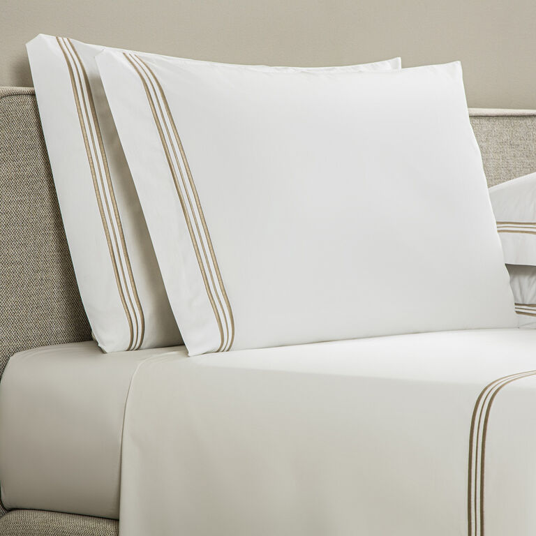 Two Frette Triplo Bourdon white cotton pillowcases and a beige-striped, three-line embroidered bedsheet are arranged on a beige upholstered bed.