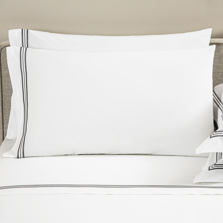 A neatly made bed with Frette’s Triplo Bourdon pillowcase set in white cotton poplin, detailed with simple black and gray three-line embroidery along the borders.