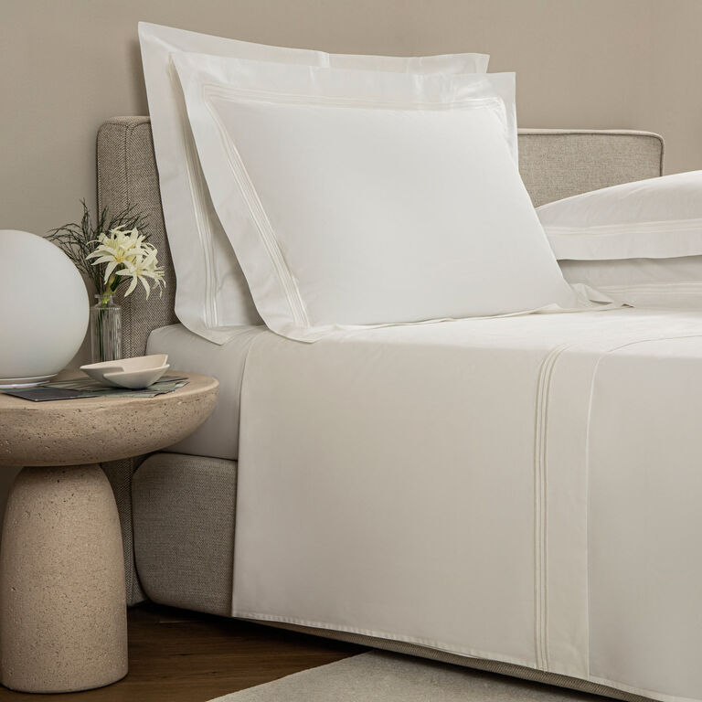 A bed is dressed in Frette’s Triplo Bourdon Sheet Set—white cotton poplin with three-line embroidery—next to a round stone nightstand displaying a glass vase of flowers, a bowl, and a spherical lamp.