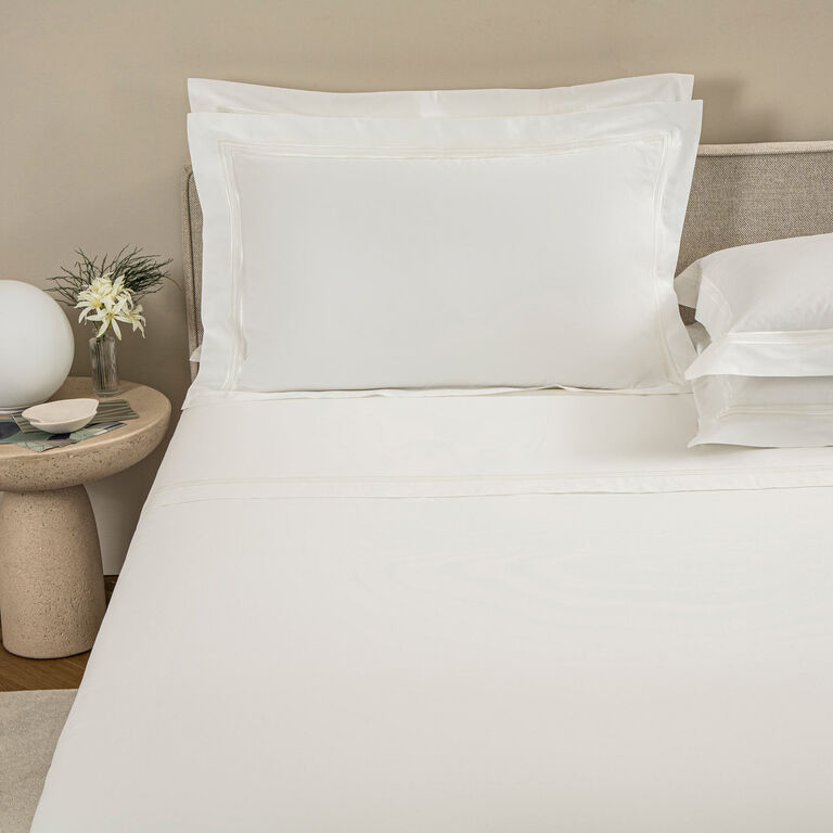 A neatly made bed with Frette's Triplo Bourdon pillowcase set, featuring subtle three-line embroidery, sits beside a round nightstand topped with a small flower vase and a round white lamp.