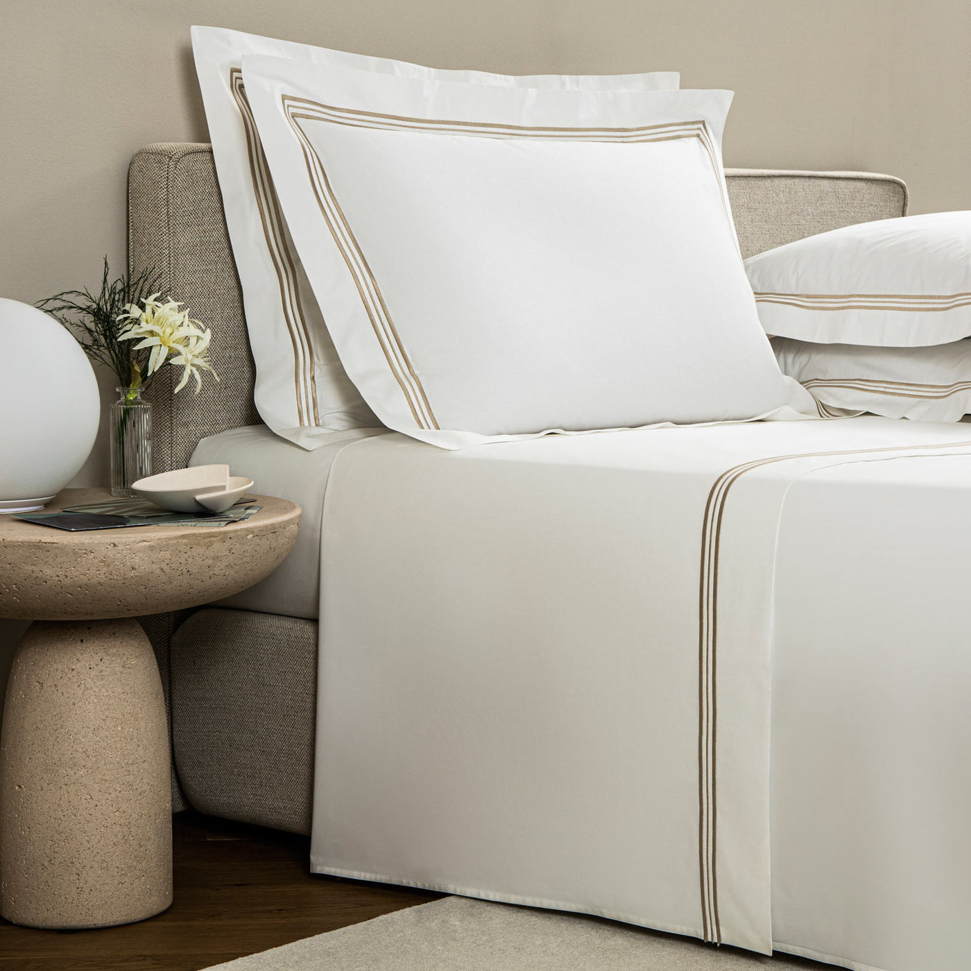 A neatly made bed showcases the Frette Triplo Bourdon Sheet Set with white sheets and pillows elegantly accented by three-line embroidery, next to a round stone side table holding a vase of flowers and a white globe lamp.