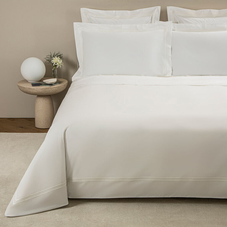 A neatly made bed features the Frette Triplo Bourdon Duvet Cover in white cotton poplin with three-line embroidery, complemented by pillows, beside a round nightstand with a globe lamp, books, and a small vase of flowers.