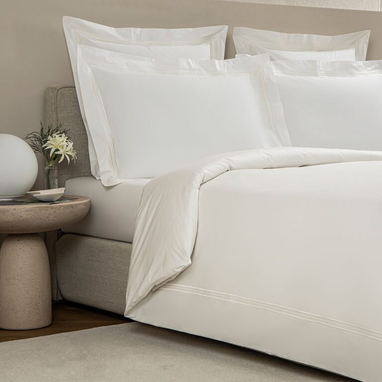 A neatly made bed with white cotton poplin pillows and the Frette Triplo Bourdon Duvet Cover featuring three-line embroidery, beside a round nightstand with a flower arrangement and a spherical lamp.