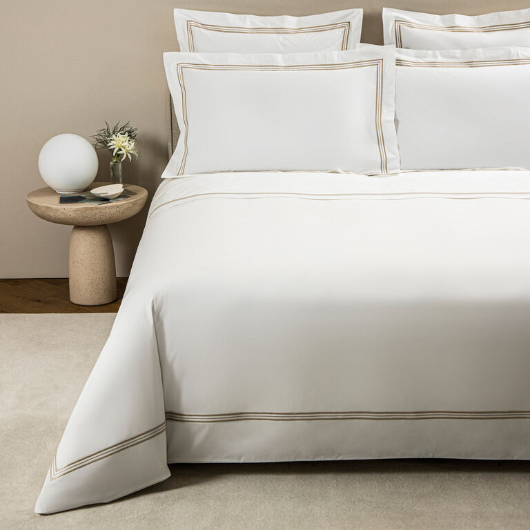 A neatly made bed with Frette's Triplo Bourdon duvet cover in white cotton poplin, adorned with elegant beige three-line embroidery, stands beside a round table holding a white lamp, a book, and a vase of flowers.
